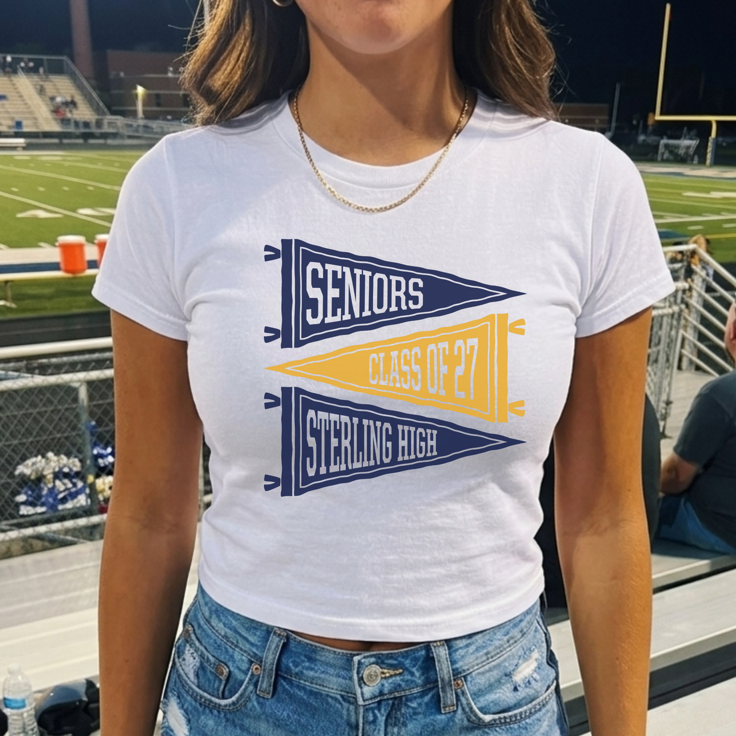 Sterling High Seniors Class of 2027 fitted white baby tee with vintage navy and gold pennant graphic shown at a football stadium.