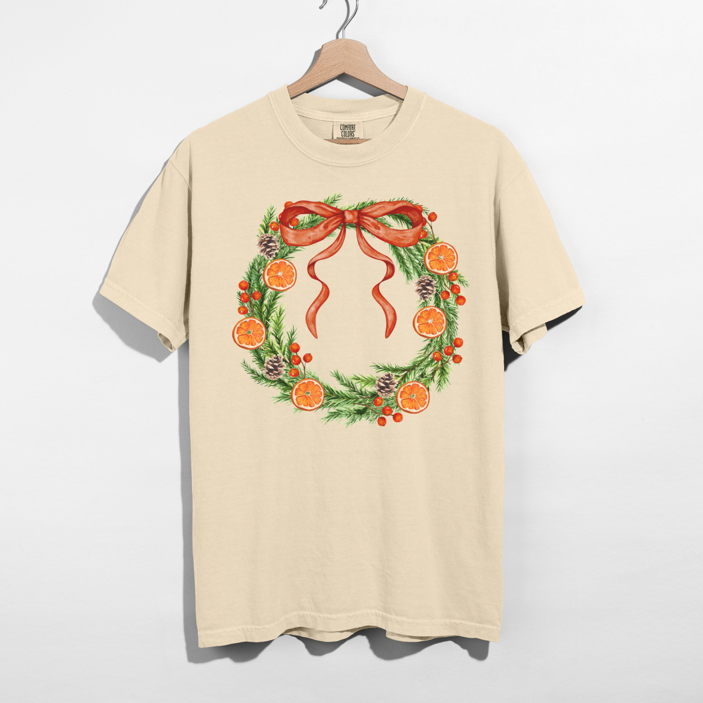 Ivory Orange Grove Christmas Wreath graphic t-shirt flat lay featuring an evergreen wreath with dried oranges, pinecones, and a red bow.