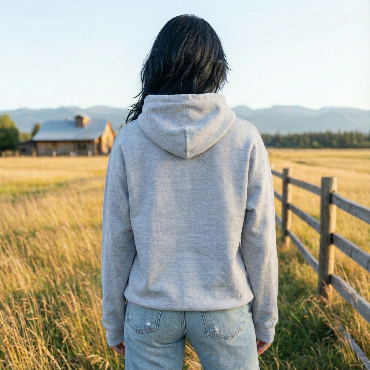 Back view of woman wearing heather grey pullover hoodie with relaxed fit, fleece hood, and distressed denim jeans in a country field
