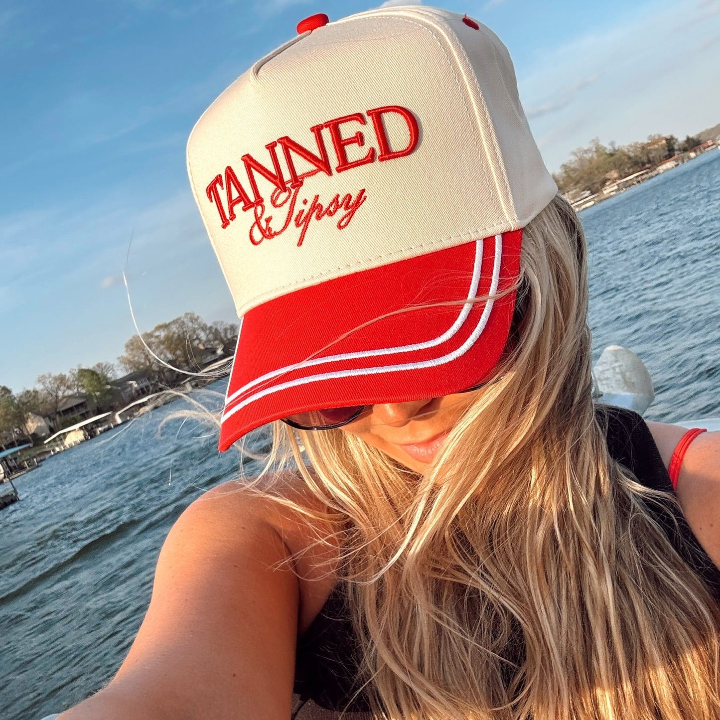 Person wearing a 'Tanned & Tipsy' cap by a body of water