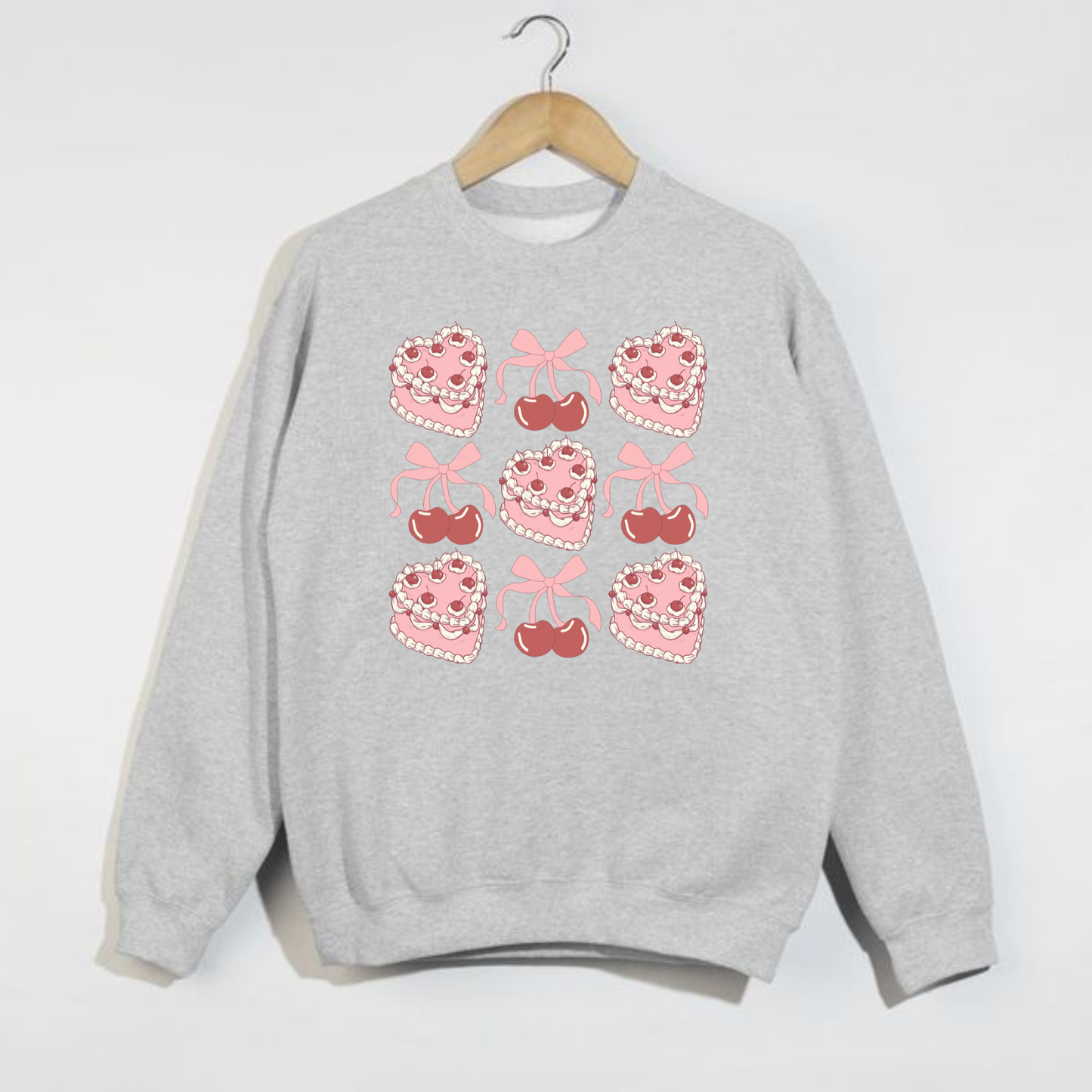 Ash grey Valentine crewneck sweatshirt on hanger featuring pink heart cakes, cherries, and bows arranged in a repeating 3x3 pattern.