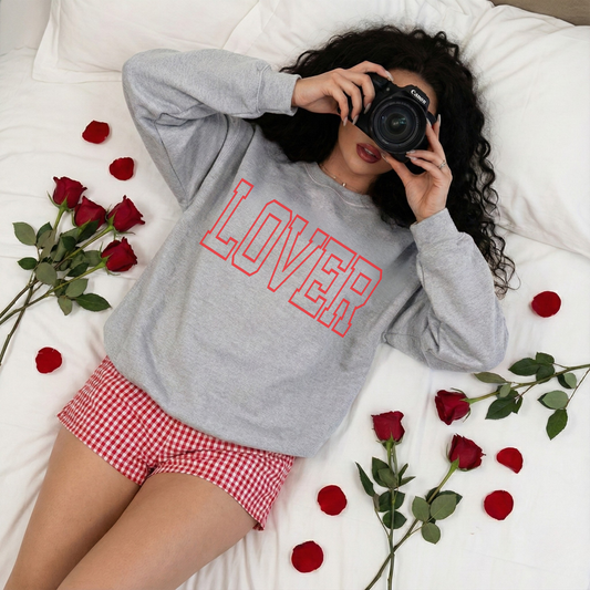 Woman lying on a bed wearing grey LOVER sweatshirt with red lettering surrounded by roses and rose petals.