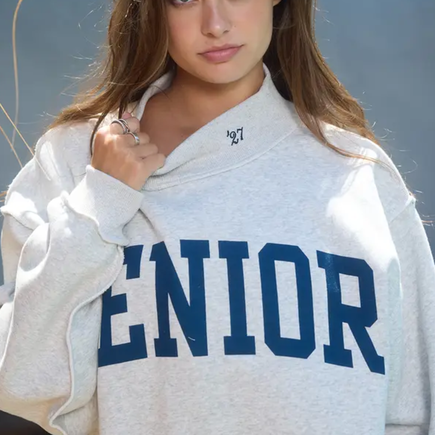 Person wearing a gray sweatshirt with 'SENIOR' printed in blue letters against a blurred outdoor background.