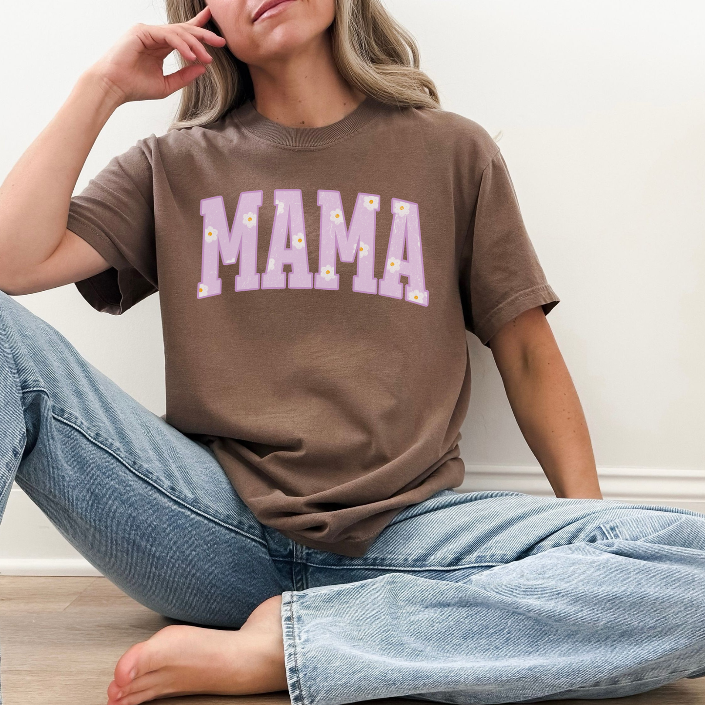 Woman wearing a brown Comfort Colors mama graphic t-shirt with purple floral collegiate lettering, casual seated fit