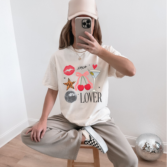 Woman sitting on stool wearing ivory Lover graphic tee with martini glass and cherry design, styled with beige pants and hat.