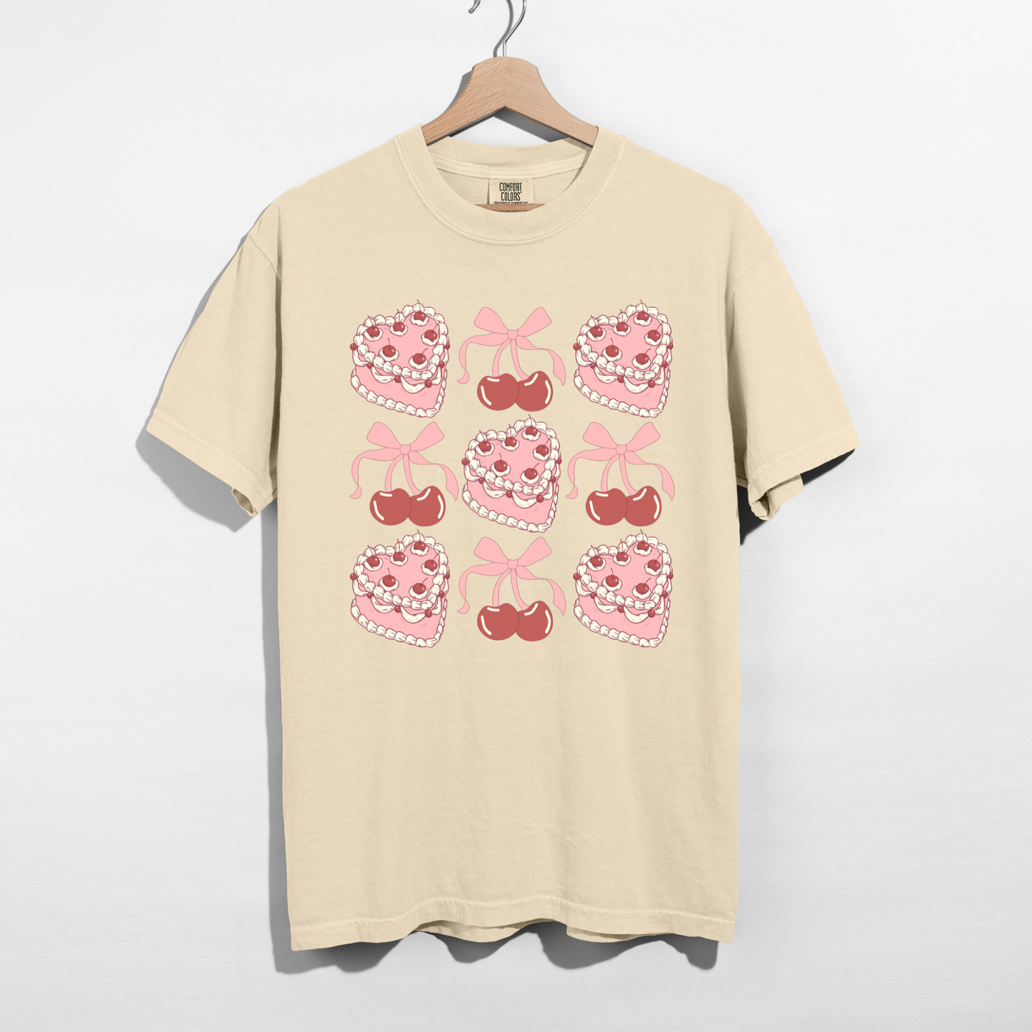Ivory Sweetheart Cherry Cake Graphic Tee featuring retro pink heart cakes, cherries, and bows displayed on a hanger.