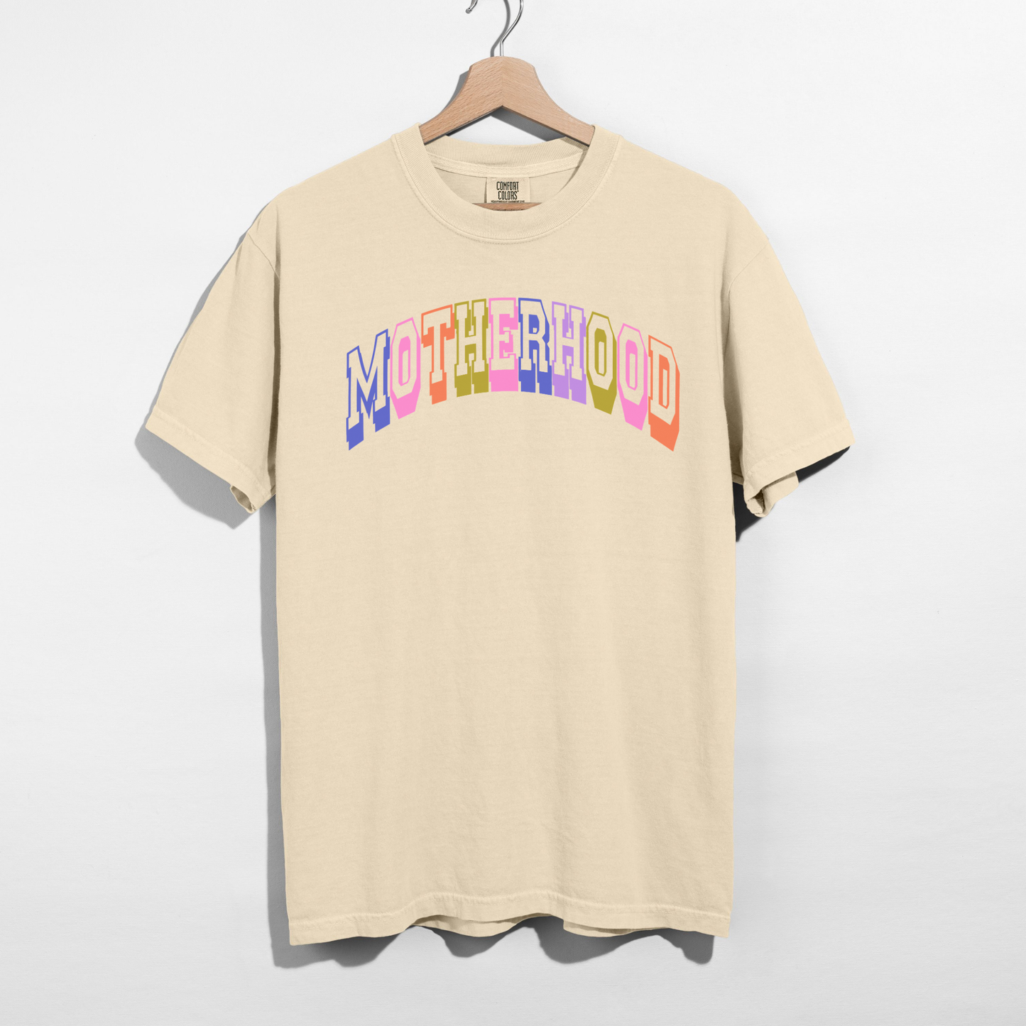 Ivory Comfort Colors 1717 retro motherhood graphic tee with pastel collegiate lettering hanging on wooden hanger.