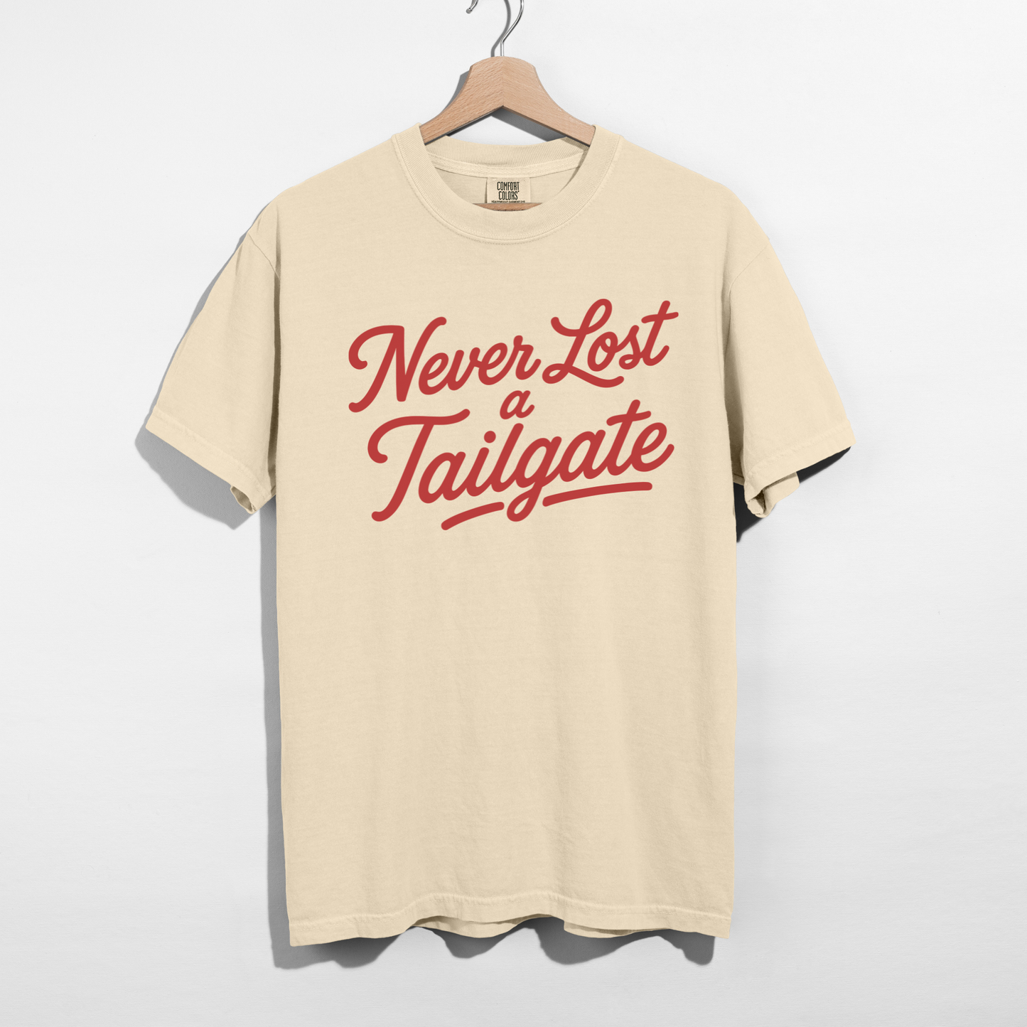 Ivory graphic T-shirt with "Never Lost a Tailgate" in bold crimson script lettering displayed on a wooden hanger.