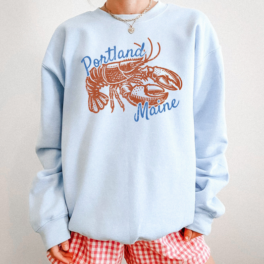 Woman wearing light blue Portland Maine sweatshirt with vintage lobster graphic, styled with red gingham shorts.