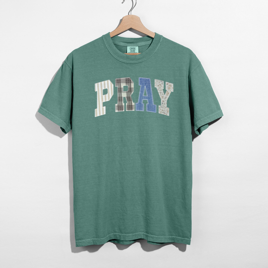 Green t-shirt with 'PRAY' printed on it, hanging on a wooden hanger against a white background.