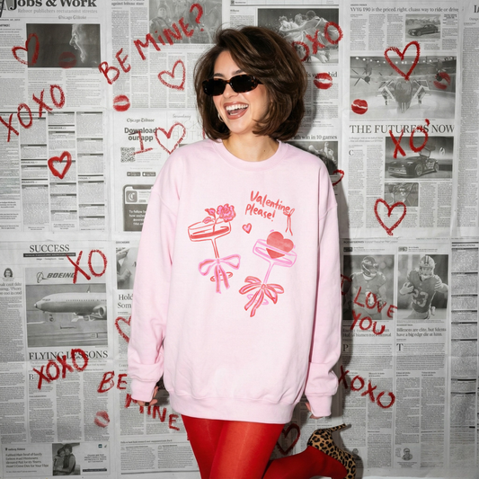 Woman wearing a light pink Valentine’s crewneck with pink and red bow designs, posing in front of a newspaper backdrop decorated with hand-drawn hearts.