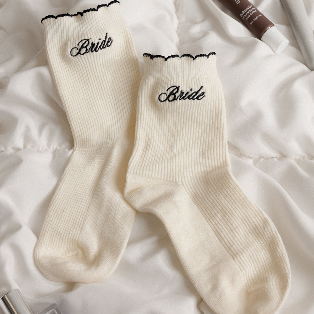 White socks with 'Bride' embroidery on a textured surface with beauty products.