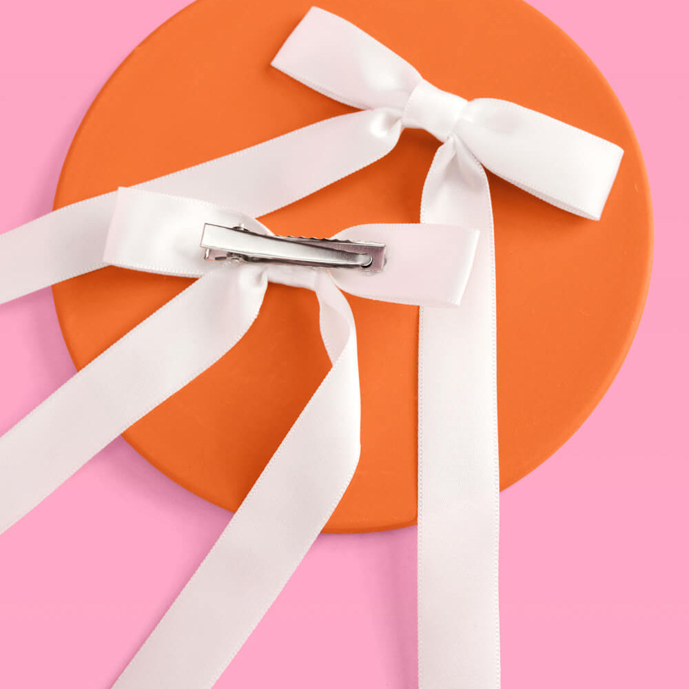 White ribbon tied in a bow with a hair clip on an orange circle against a pink background