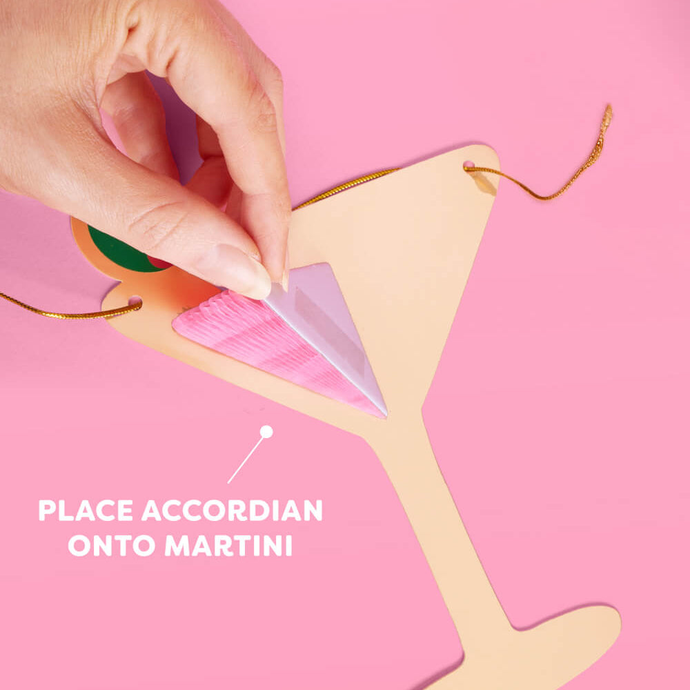 Hand placing an accordion onto a martini glass cutout on a pink background