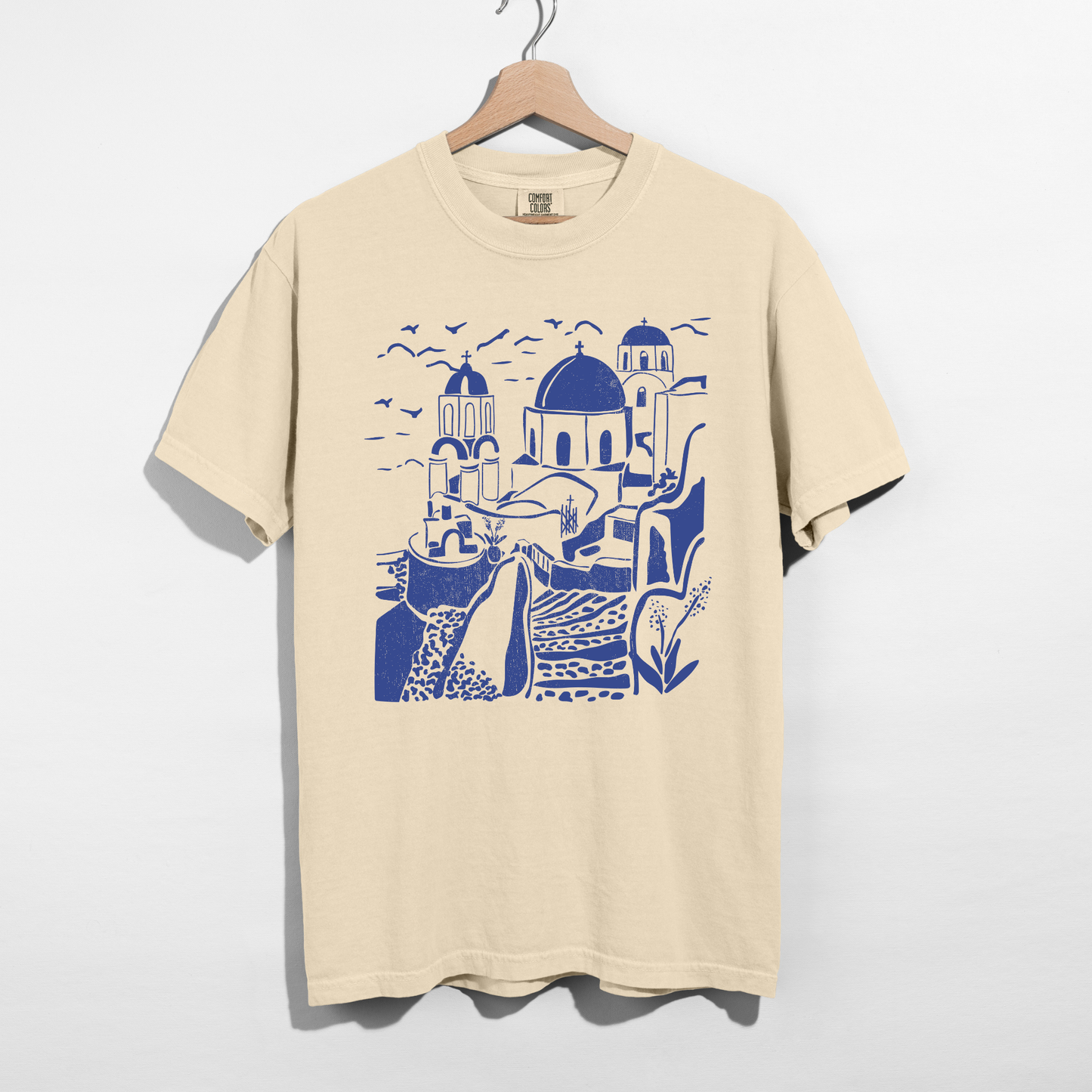 Comfort Colors cream Santorini Greece graphic tee on wooden hanger with blue Mediterranean illustration against white background.