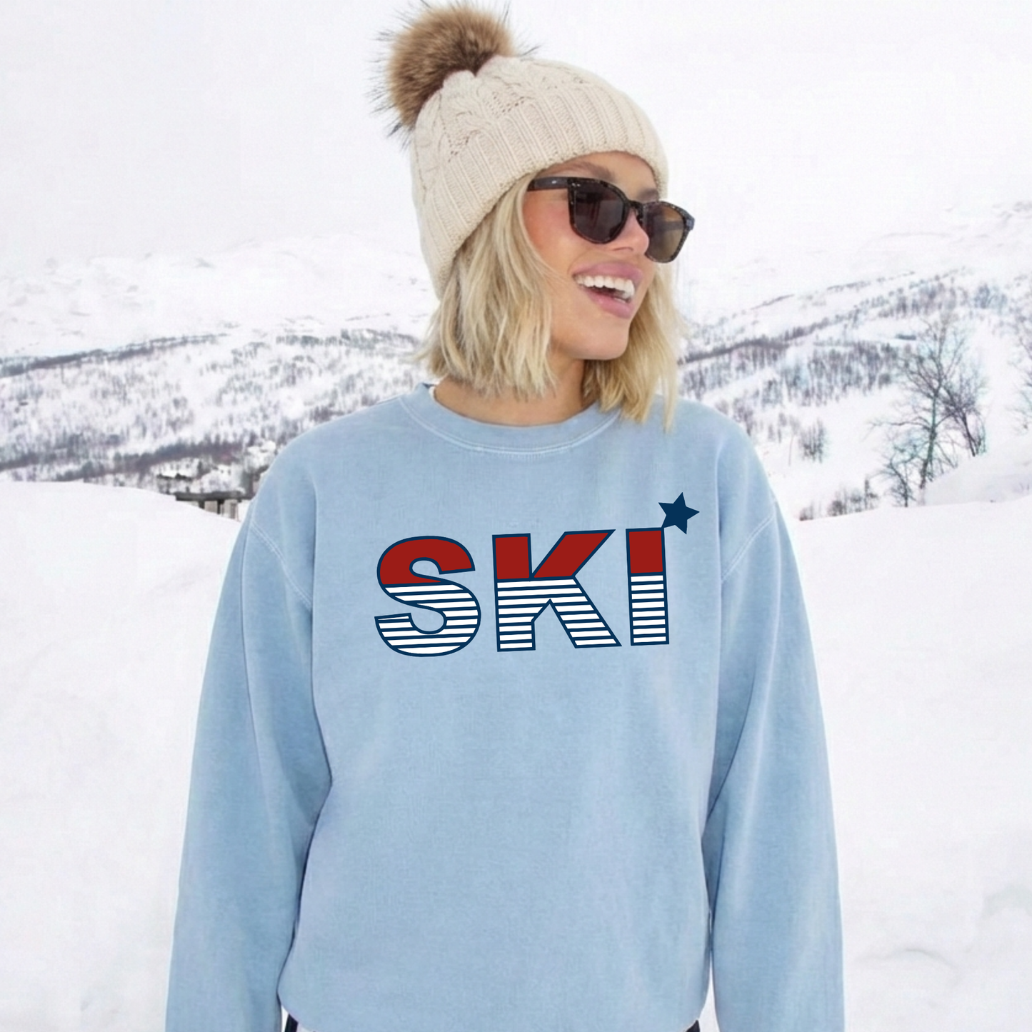 Woman wearing a light blue ski sweatshirt with retro red white and blue SKI graphic while standing in a winter mountain landscape.