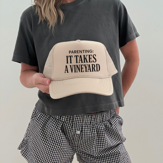 Cream trucker hat with “Parenting: It Takes a Vineyard” text held by model wearing casual outfit