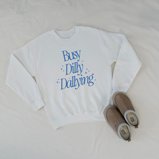 White sweatshirt with blue text 'Busy Dilly Dallying' and brown slippers on a light gray background