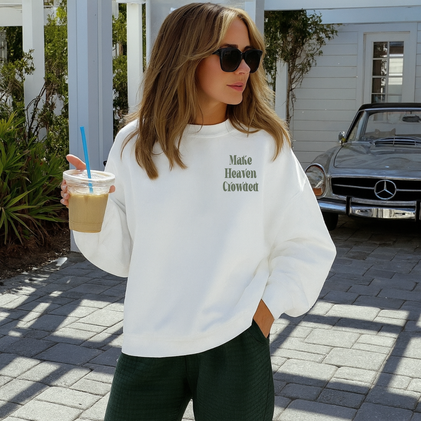 “Woman wearing white crewneck with small green ‘Make Heaven Crowded’ design on the front, holding iced coffee outdoors.”