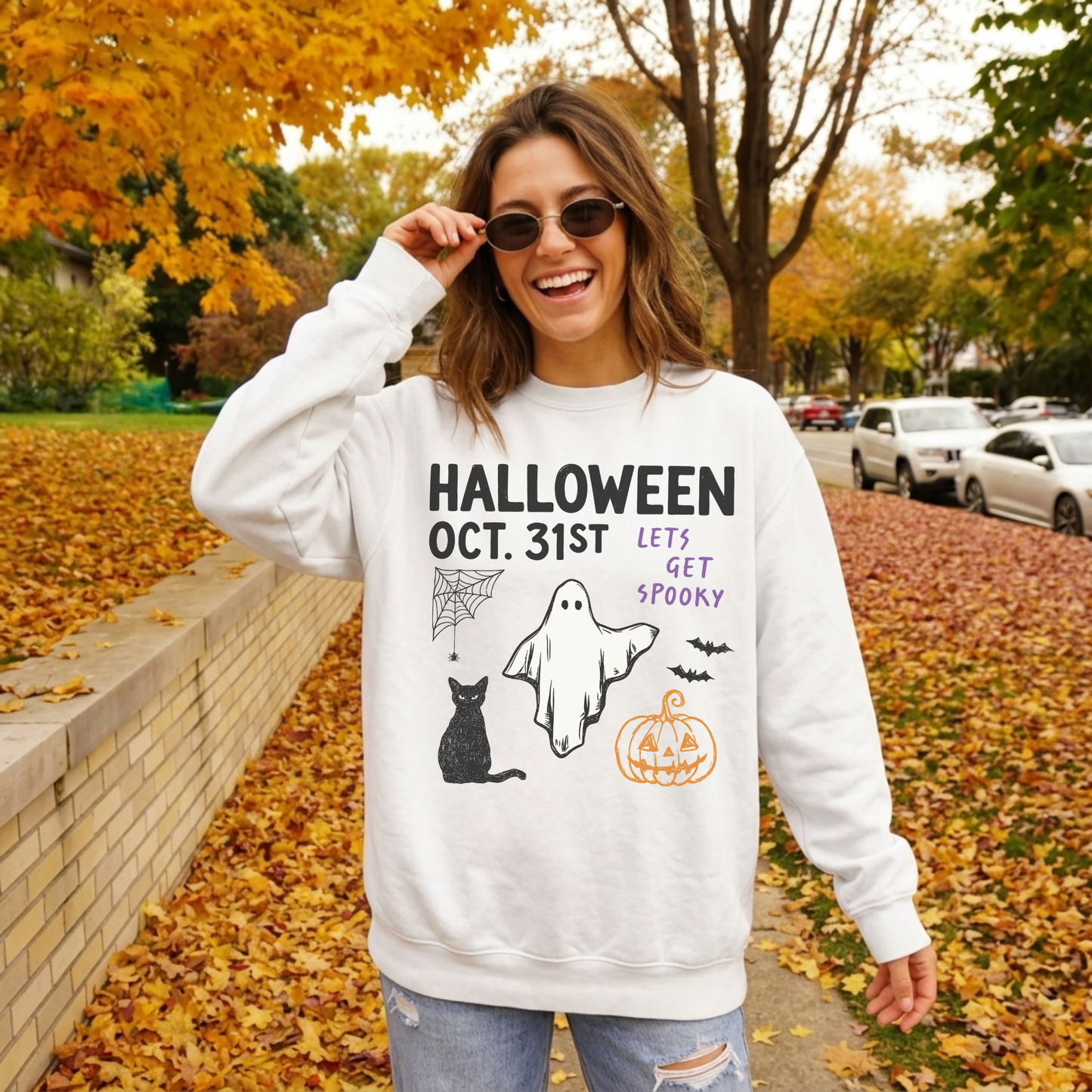 Woman wearing a white Halloween Oct. 31st ghost graphic sweatshirt with black cat, pumpkin, bats and spooky Halloween design while walking through colorful fall leaves.