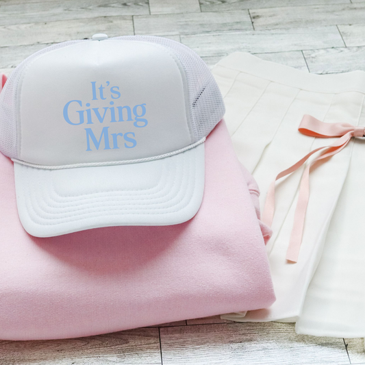 White foam front trucker hat with “It’s Giving Mrs” text styled with pink sweatshirt and bridal outfit