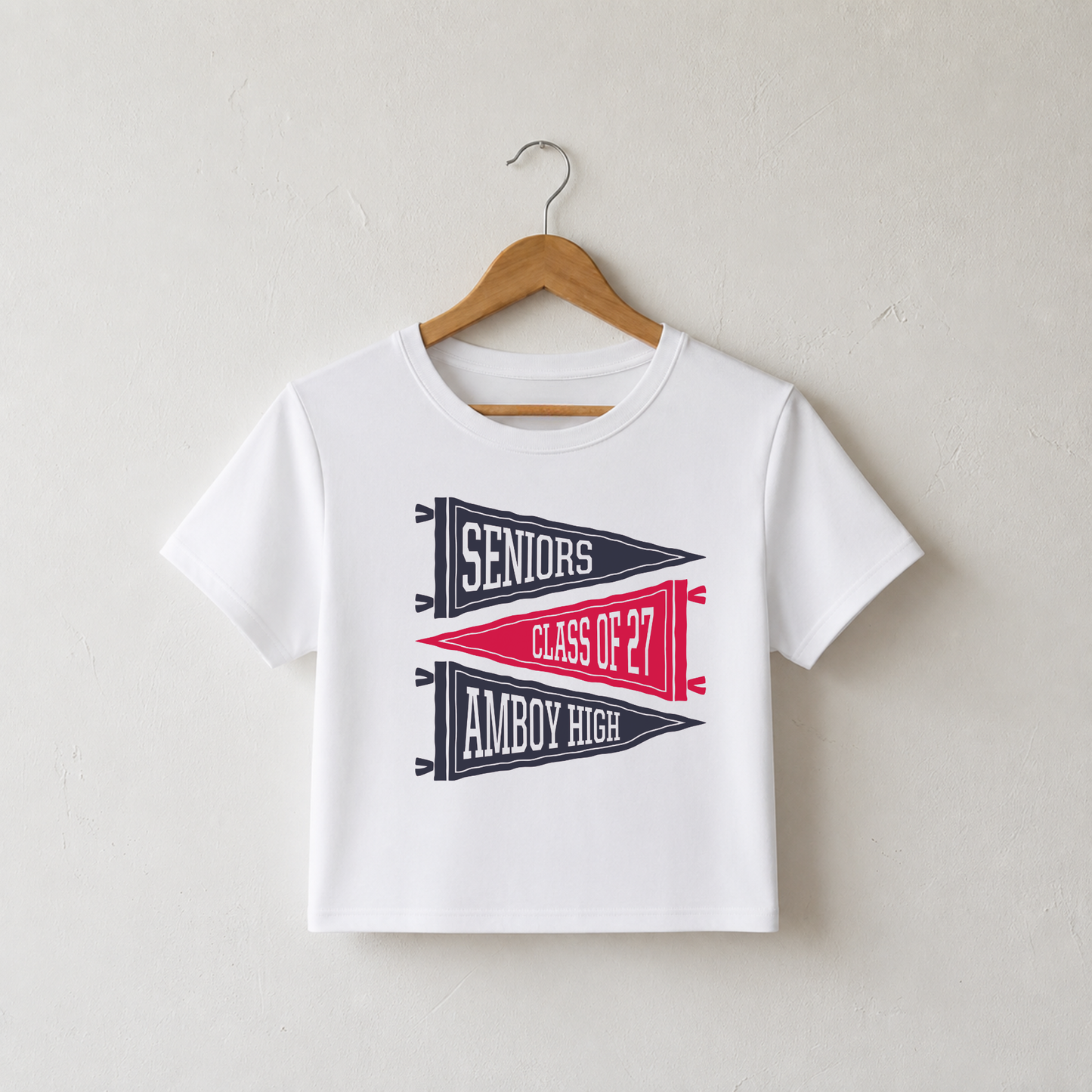 White women's fitted baby tee featuring a vintage navy and red Amboy High Seniors Class of 2027 pennant graphic hanging on a wooden hanger.
