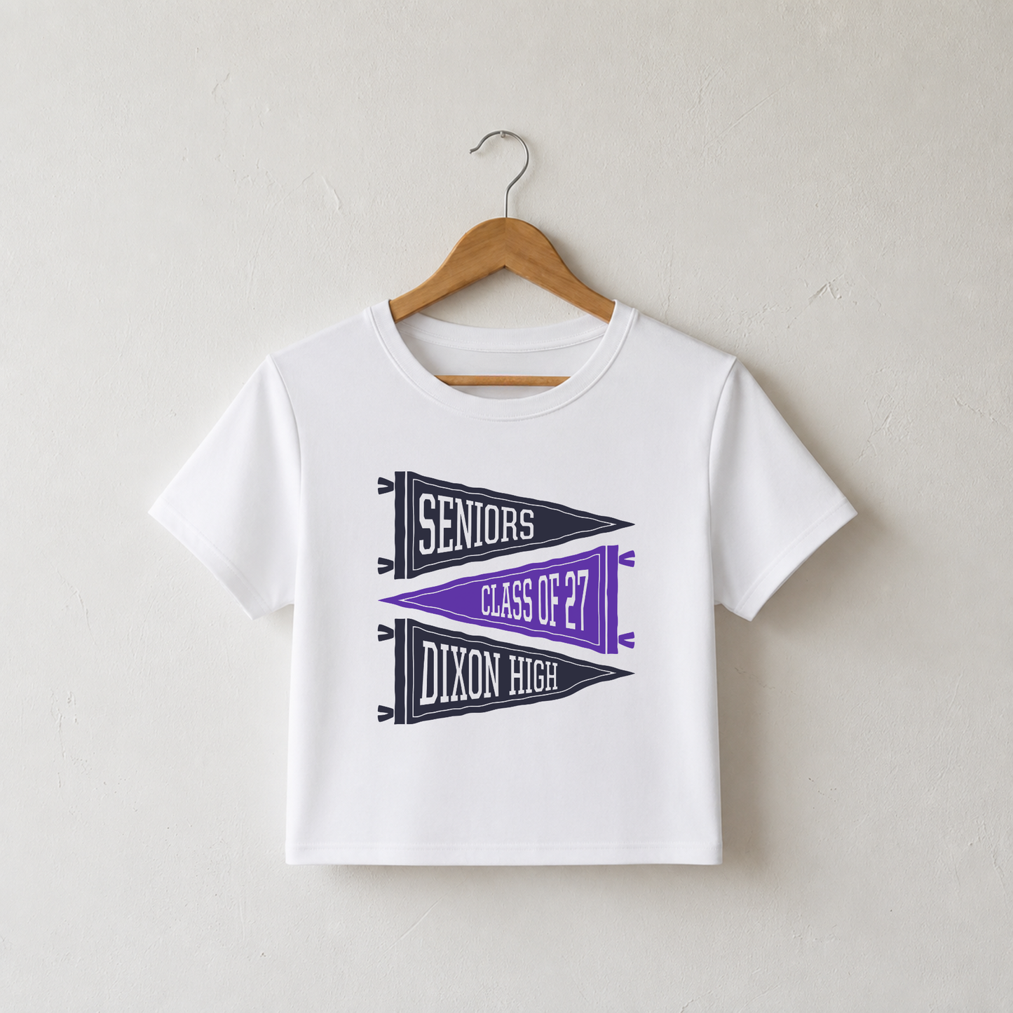 White fitted baby tee featuring Dixon High Seniors Class of 2027 purple pennant graphic hanging on wooden hanger.