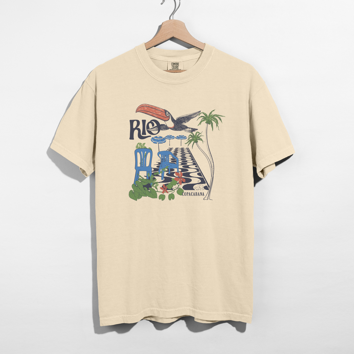 Ivory Comfort Colors Rio Copacabana graphic tee on wooden hanger against white background.
