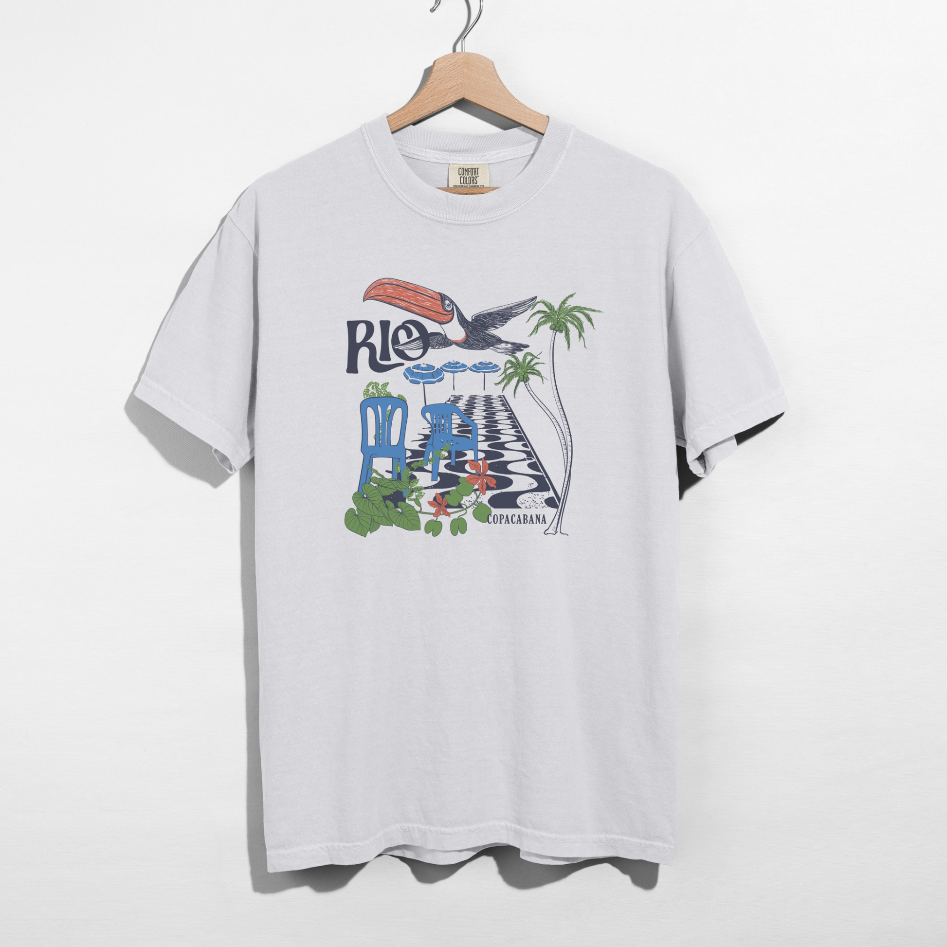 White Comfort Colors Rio Copacabana graphic tee on wooden hanger against white background.
