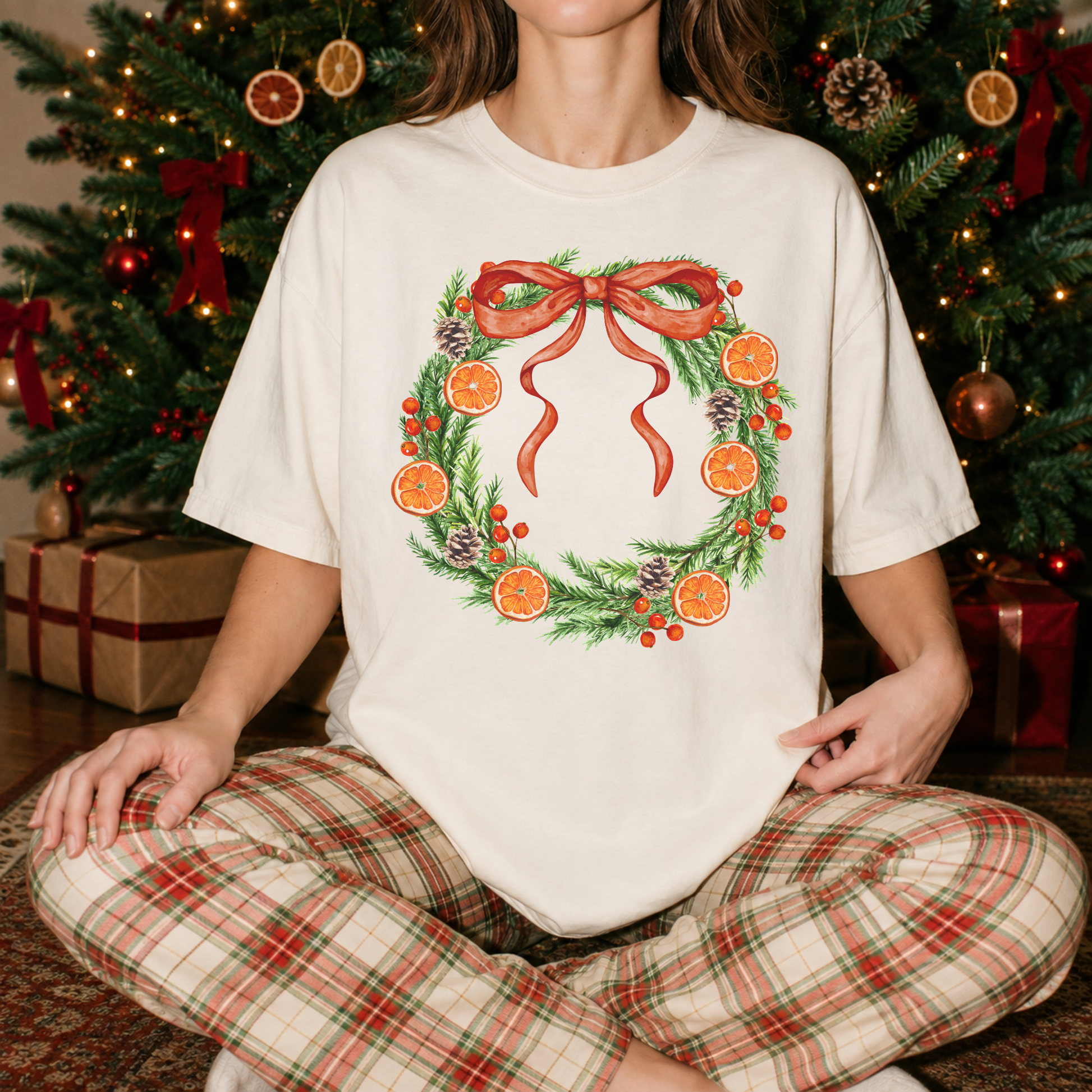 Woman wearing an ivory Orange Grove Christmas Wreath Christmas graphic tee with plaid pajama pants in front of a decorated Christmas tree.