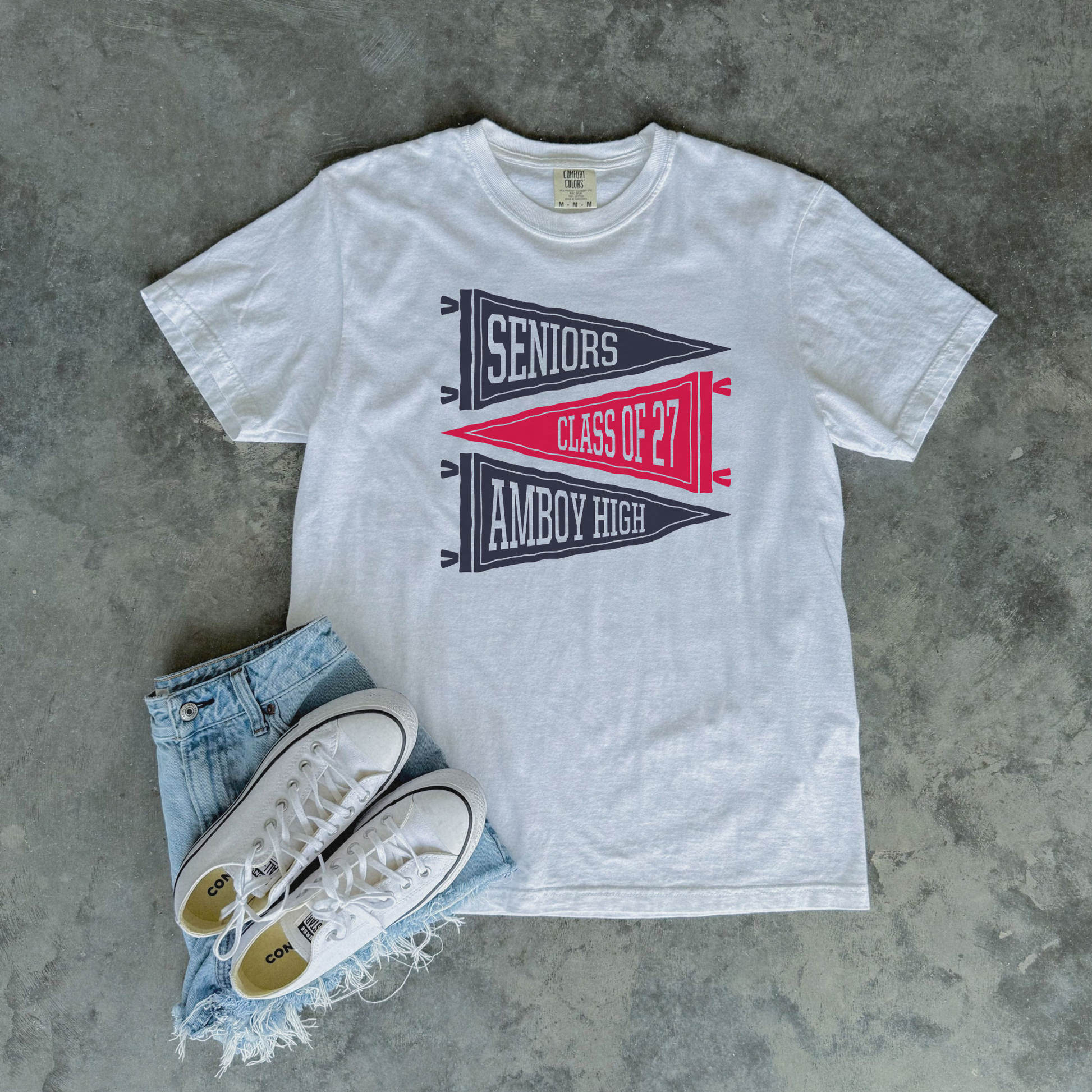 White Amboy High School Seniors Class of 2027 graphic t-shirt featuring vintage pennant flags styled with denim shorts and white sneakers.