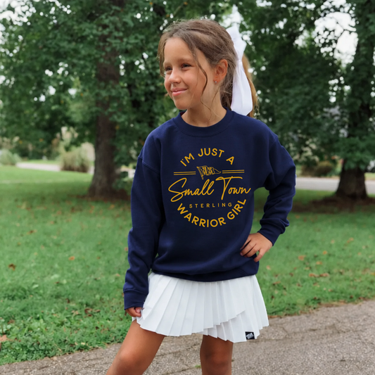 Youth girl wearing a navy Small Town Warrior Girl Sterling football crewneck sweatshirt with a white tennis skirt while posing outdoors.