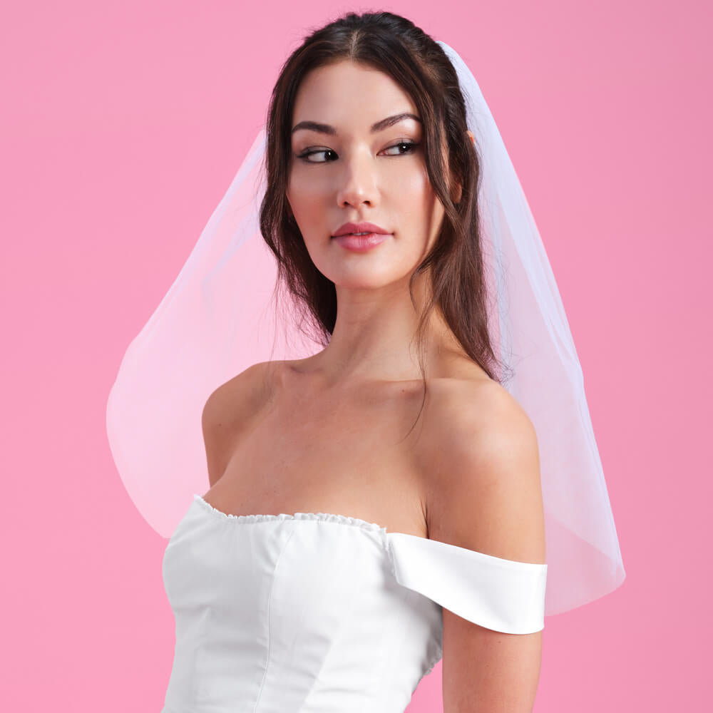 Woman wearing a white off-shoulder dress with a sheer veil against a pink background