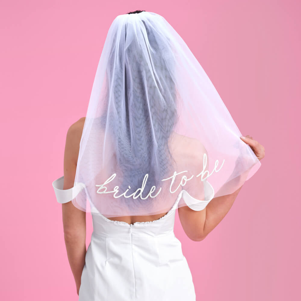 Person wearing a 'bride to be' veil against a pink background