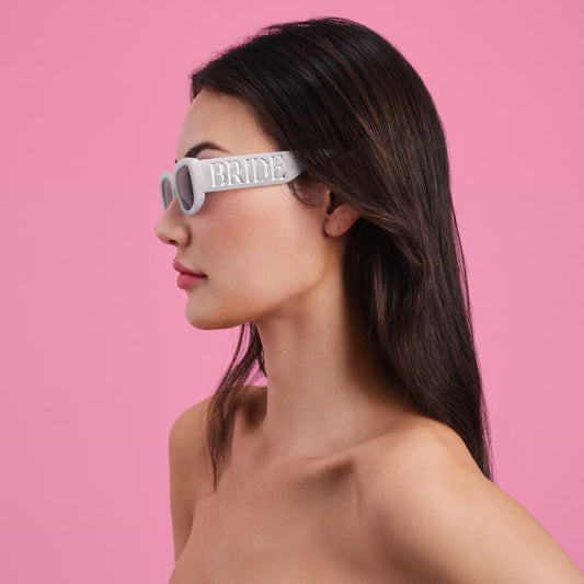 Woman wearing 'BRIDE' sunglasses with a sequin top against a pink background