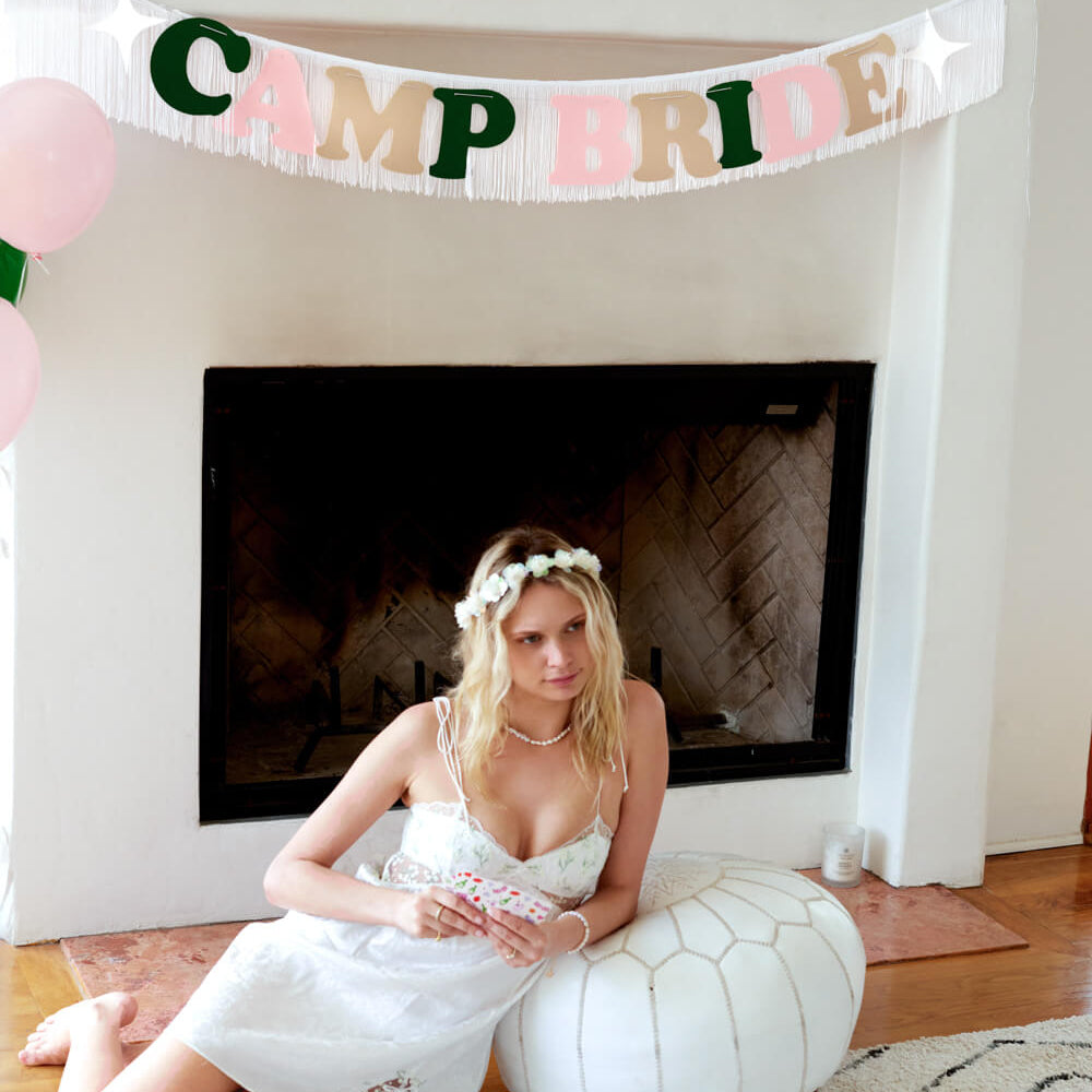 Woman in a white dress sitting on a rug with a 'Camp Bride' banner above her.