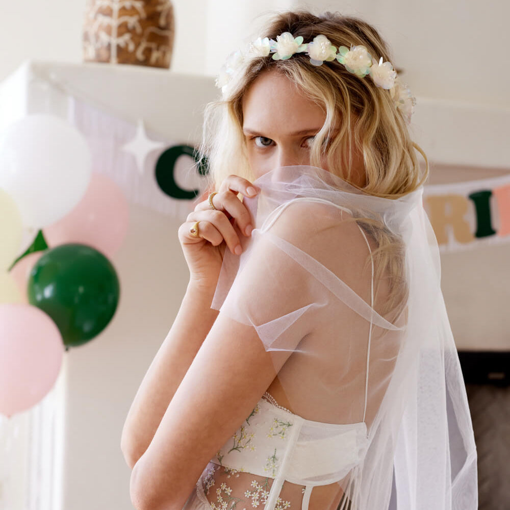 Woman wearing a white dress with sheer overlay and floral headband in a decorated room.