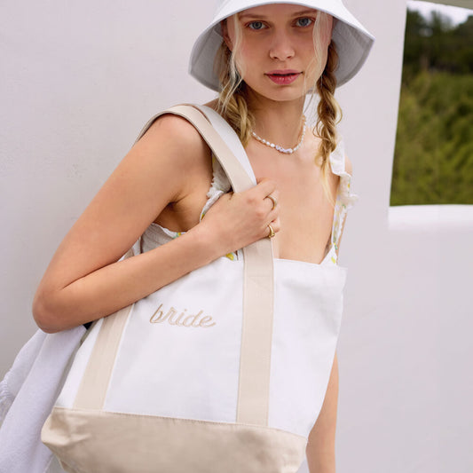Woman wearing a white 'bride' hat and holding a matching tote bag outdoors.