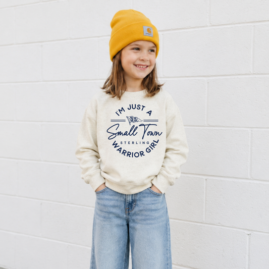 Youth girl wearing a natural heather crewneck sweatshirt featuring a navy I'm Just a Small Town Warrior Girl Sterling football graphic with light wash jeans and a gold beanie.
