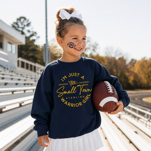 Toddler girl wearing a navy Small Town Warrior Girl football sweatshirt with a white skirt while holding a football in stadium bleachers.