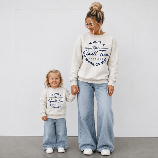 Woman and toddler wearing matching natural heather I'm Just a Small Town Warrior Girl Sterling football crewneck sweatshirts with light wash jeans for game day.