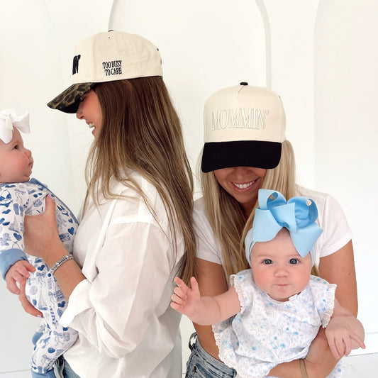 Two women holding babies, both wearing hats with text, against a white background
