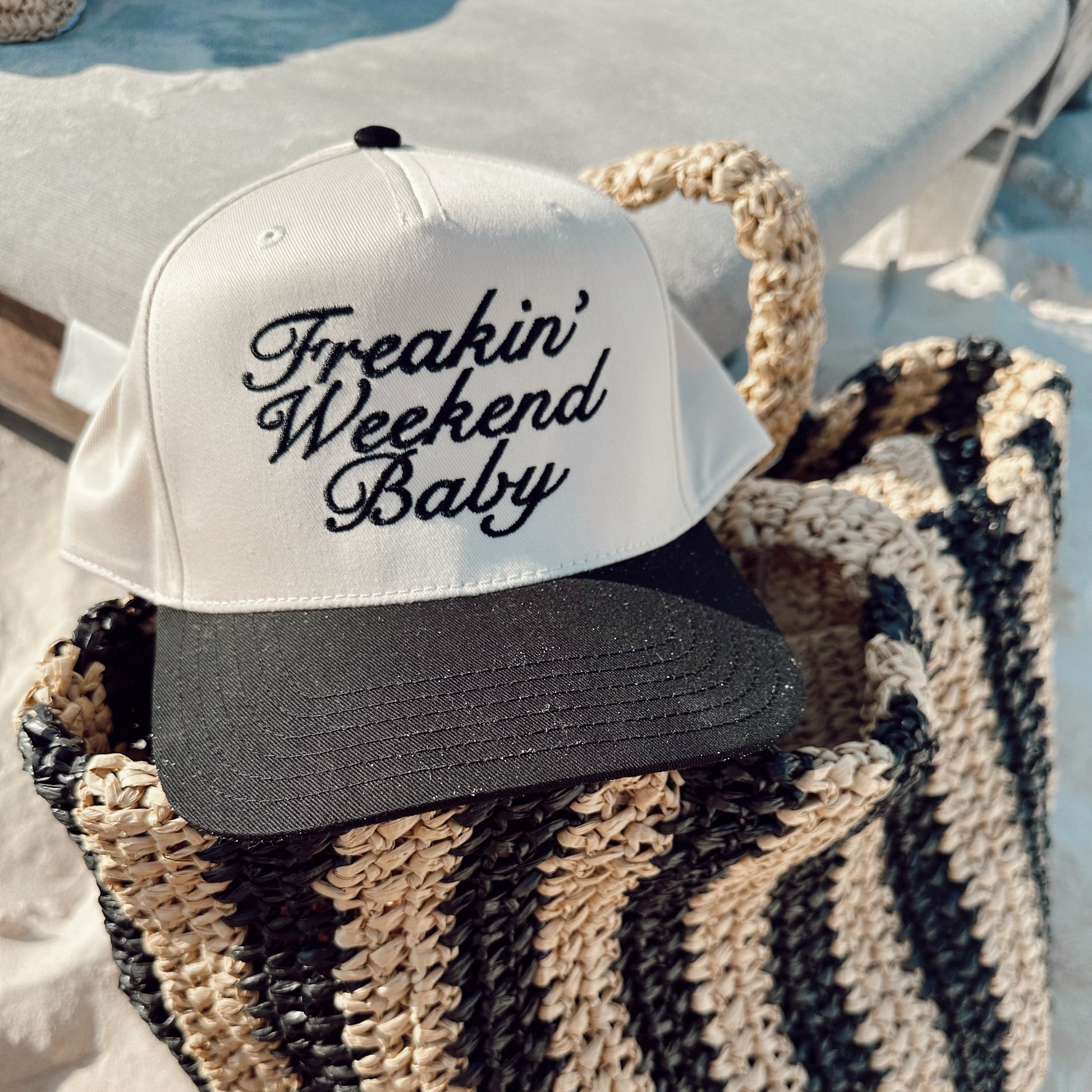 Cap with 'Freakin' Weekend Baby' text on a woven bag at the beach.