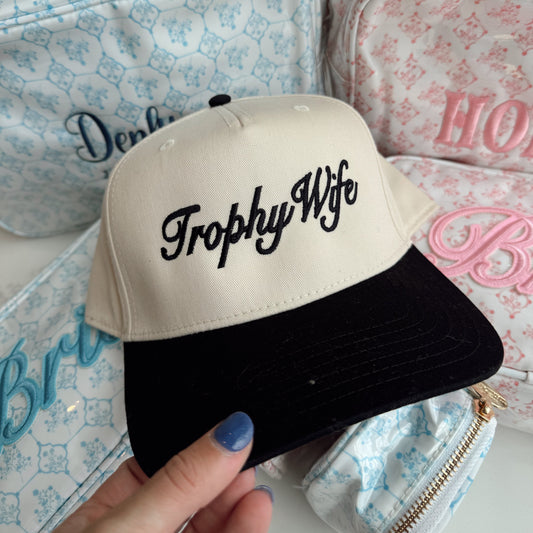 Cap with 'Trophy Wife' text held in front of patterned bags