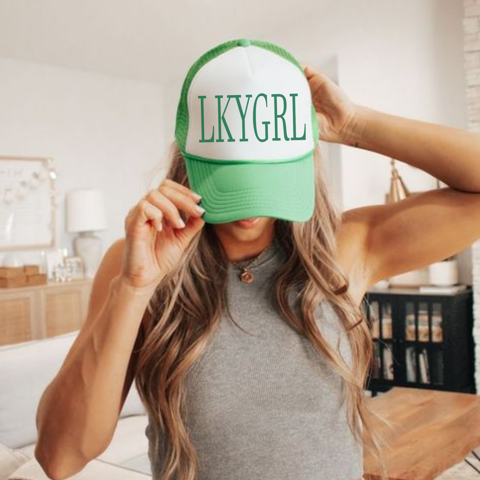 A woman wearing a green and white trucker hat with 'LKYGRL' printed on it.