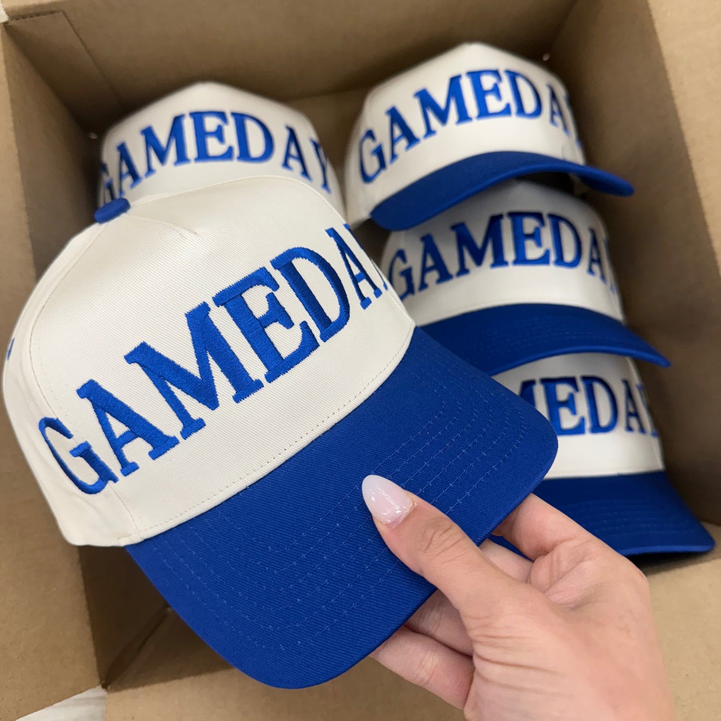 Three white caps with blue brims and 'GAMEDAY' text in a cardboard box.