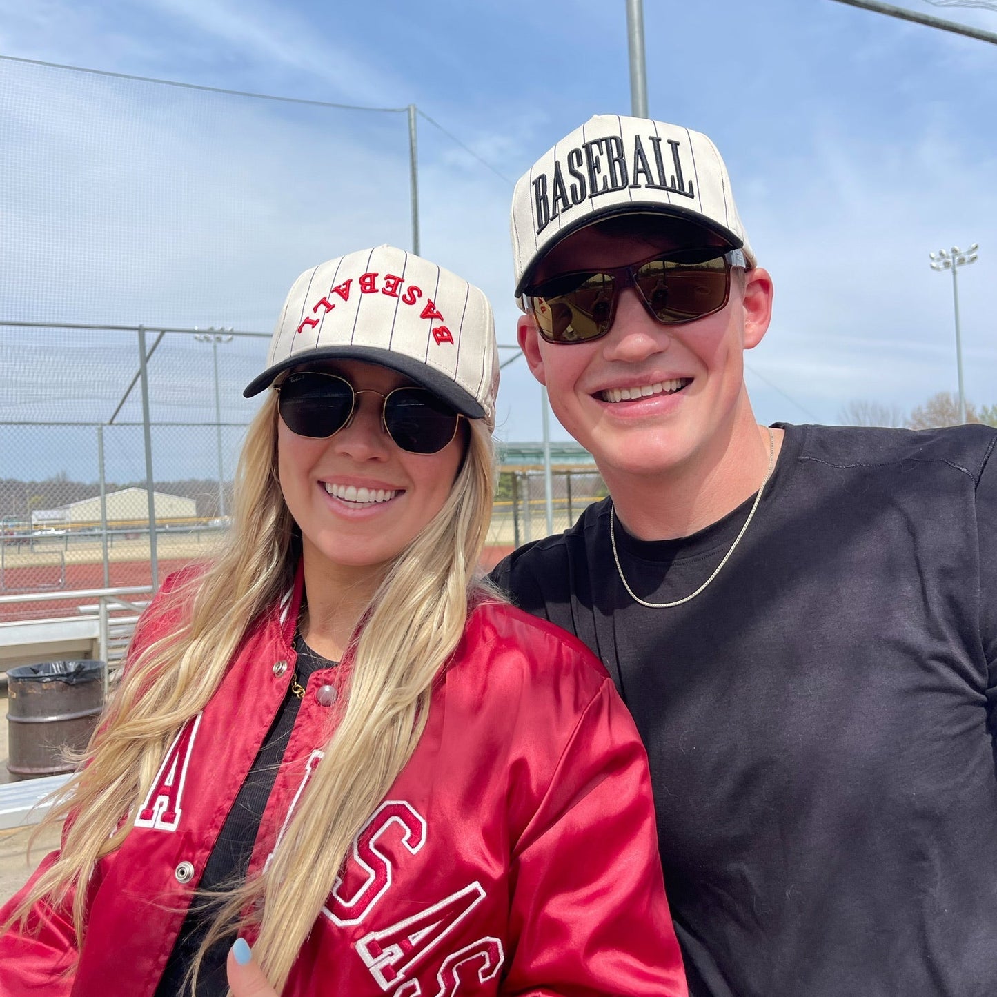 Two people wearing baseball-themed hats and sunglasses outdoors with a clear sky.