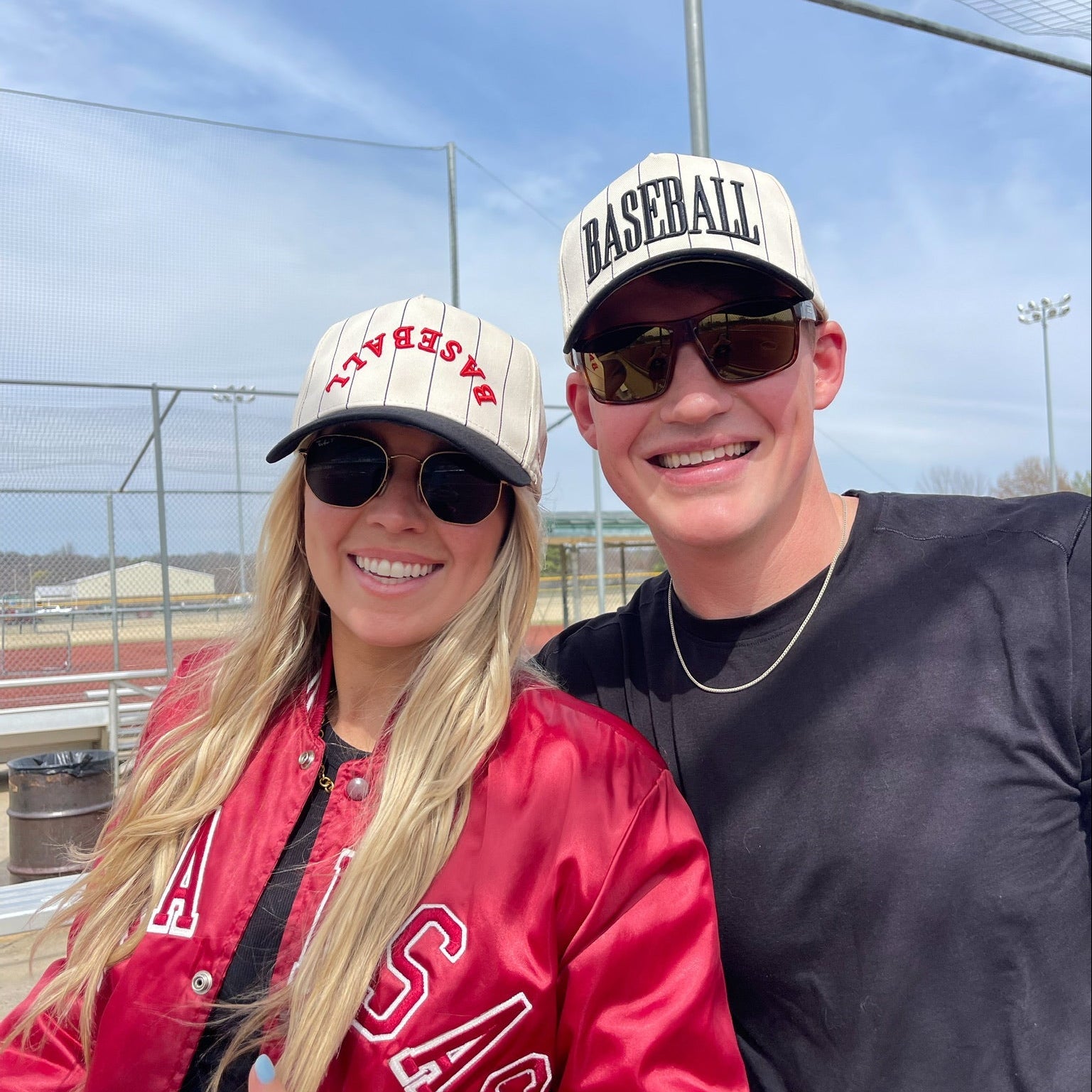 Two people wearing baseball-themed hats and sunglasses outdoors with a clear sky.