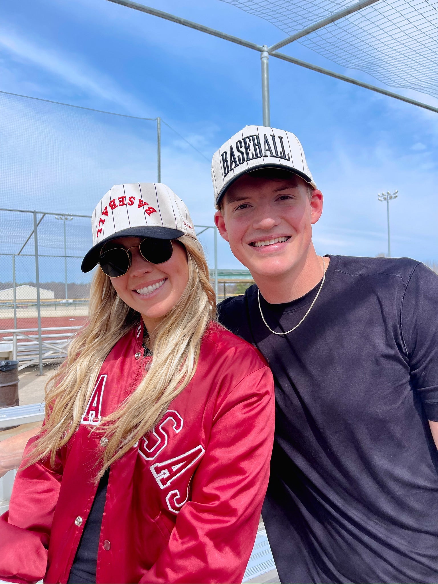 Two people wearing baseball caps and posing outdoors with a clear blue sky.