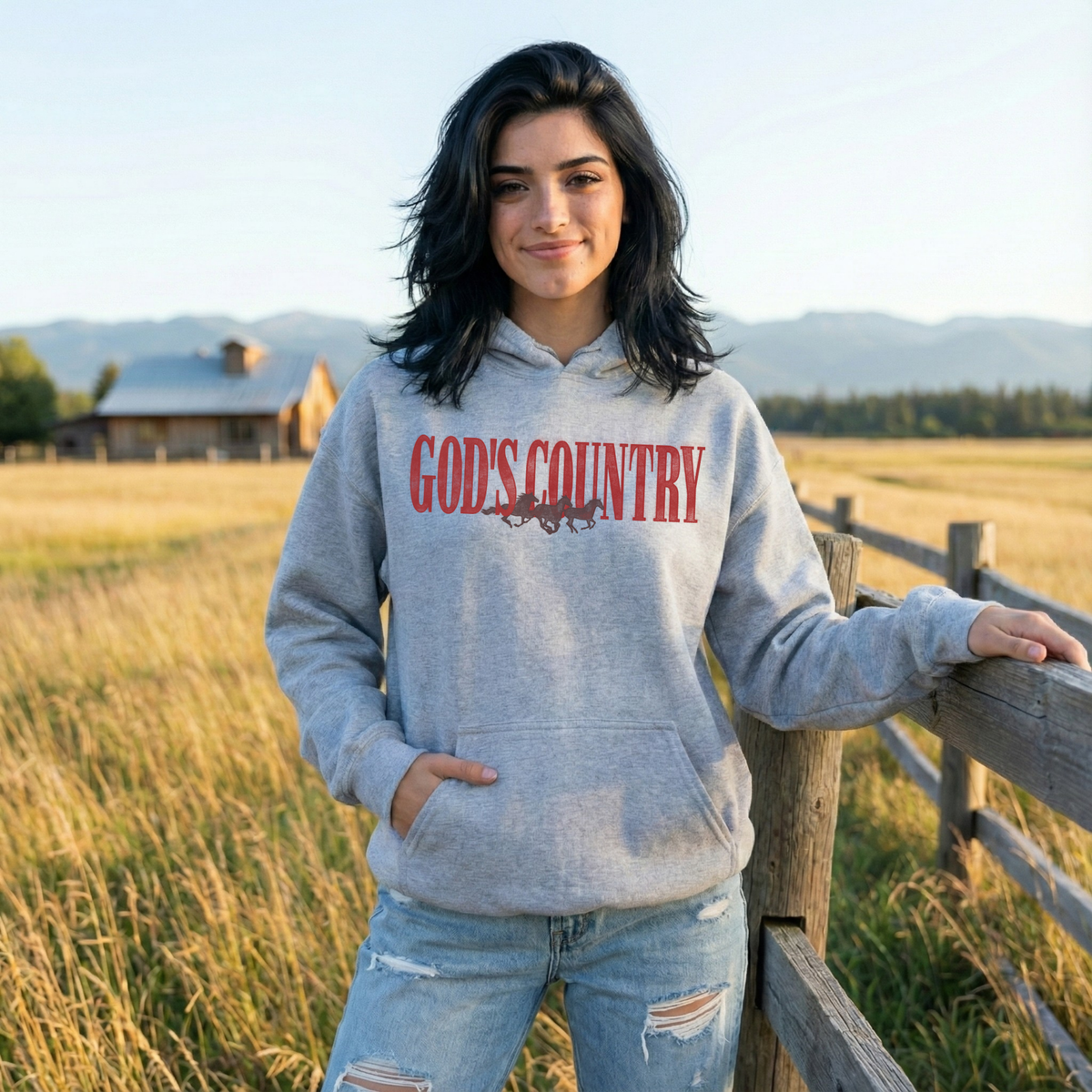 Woman wearing heather grey God's Country western graphic hoodie with vintage red lettering and running horses, styled with distressed jeans beside a wooden ranch fence